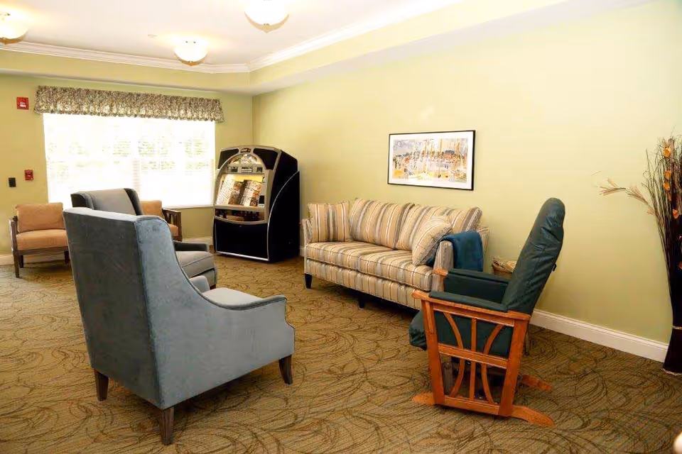 A cozy living room with a striped sofa, two blue armchairs, and a wooden rocking chair with a green cushion. There is a large window with a floral valance, a framed picture on the wall, a jukebox in the corner, and a tall vase with decorative branches on the right side.