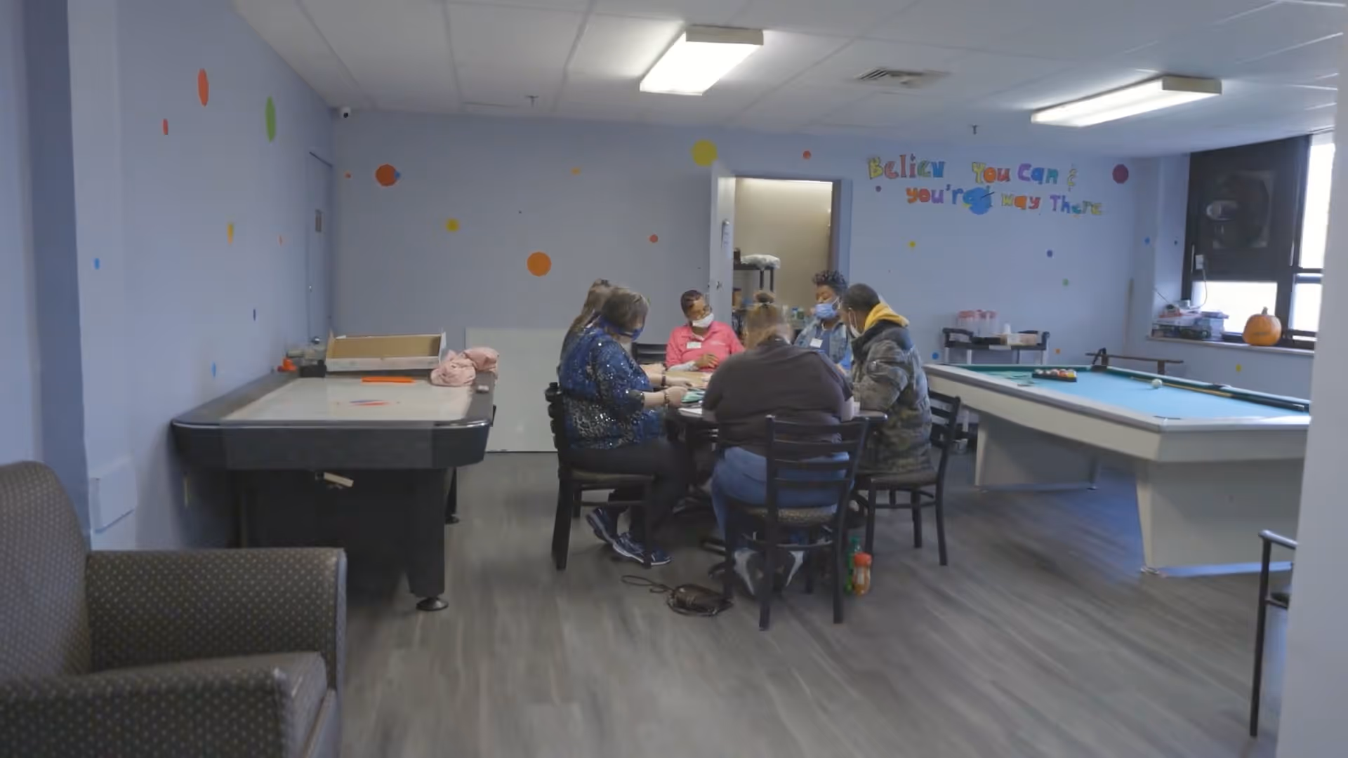 A group of five people sitting around a table playing a game in a recreational room with an air hockey table on the left and a pool table on the right. The walls are light blue with colorful dots and a motivational phrase that reads 'Believe you can & you're halfway there.' There is a window on the right side letting in natural light.