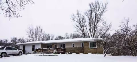 Snow-covered, single-story senior living facility with a wooden front porch and a parked car.