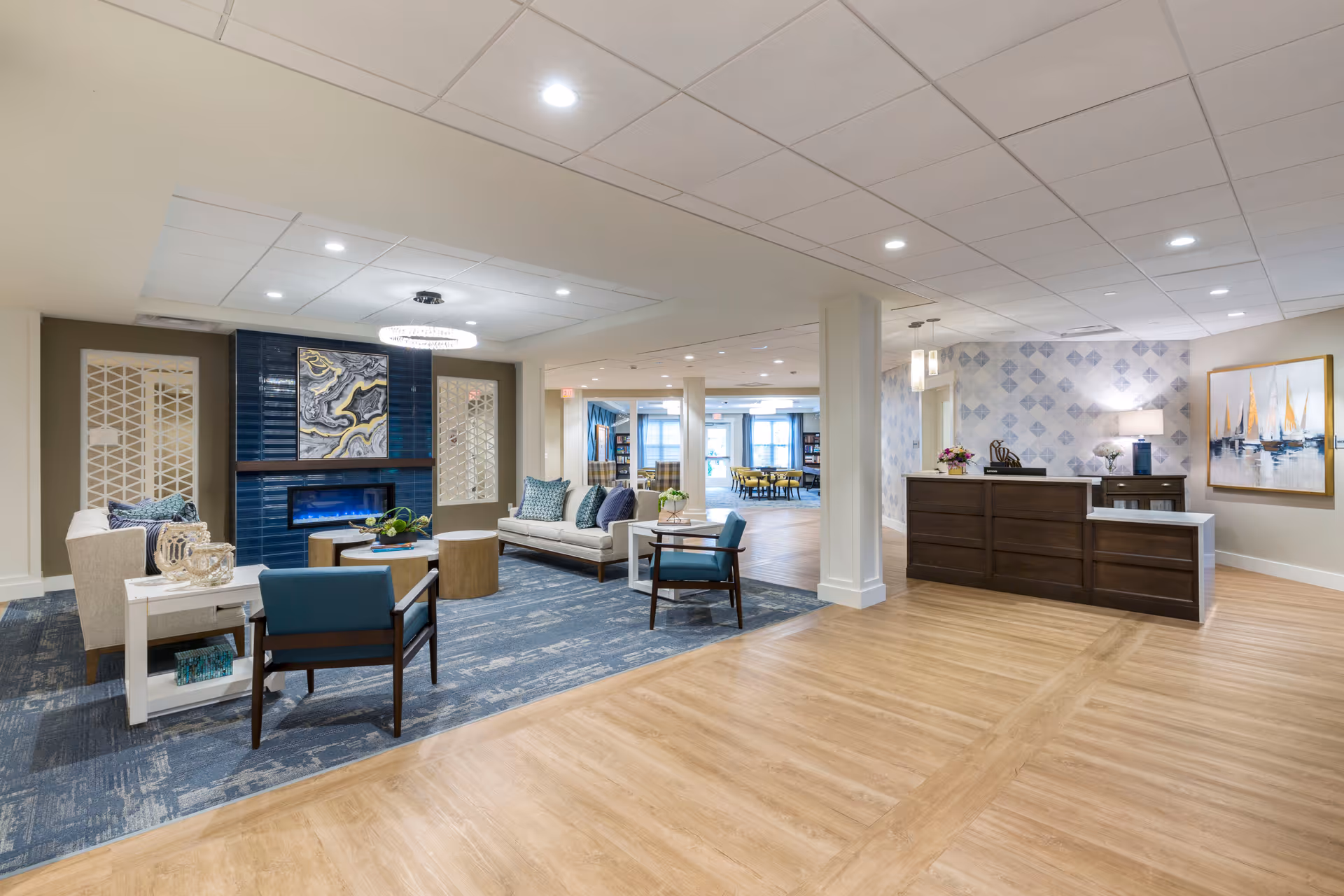 A spacious and modern senior living facility common area featuring a seating area with sofas, armchairs, and a coffee table on a blue carpet. There is a decorative fireplace with abstract art above it. To the right, there is a reception desk with a lamp, flowers, and artwork on the wall. The floor is a combination of wood and carpet, and the ceiling has recessed lighting.