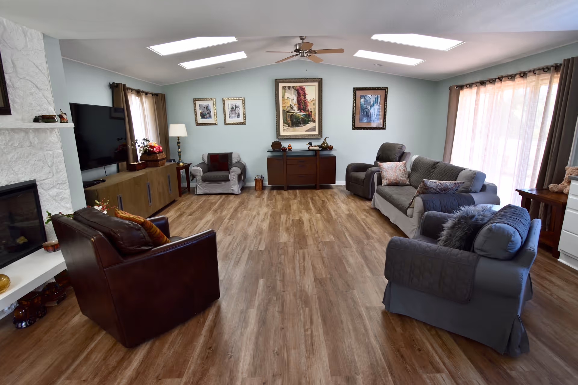 A spacious and well-lit living room with wooden flooring, multiple armchairs and sofas arranged around the perimeter, a flat-screen TV mounted on the wall, a white stone fireplace, framed artwork on the walls, large windows with curtains allowing natural light, and a ceiling fan overhead.