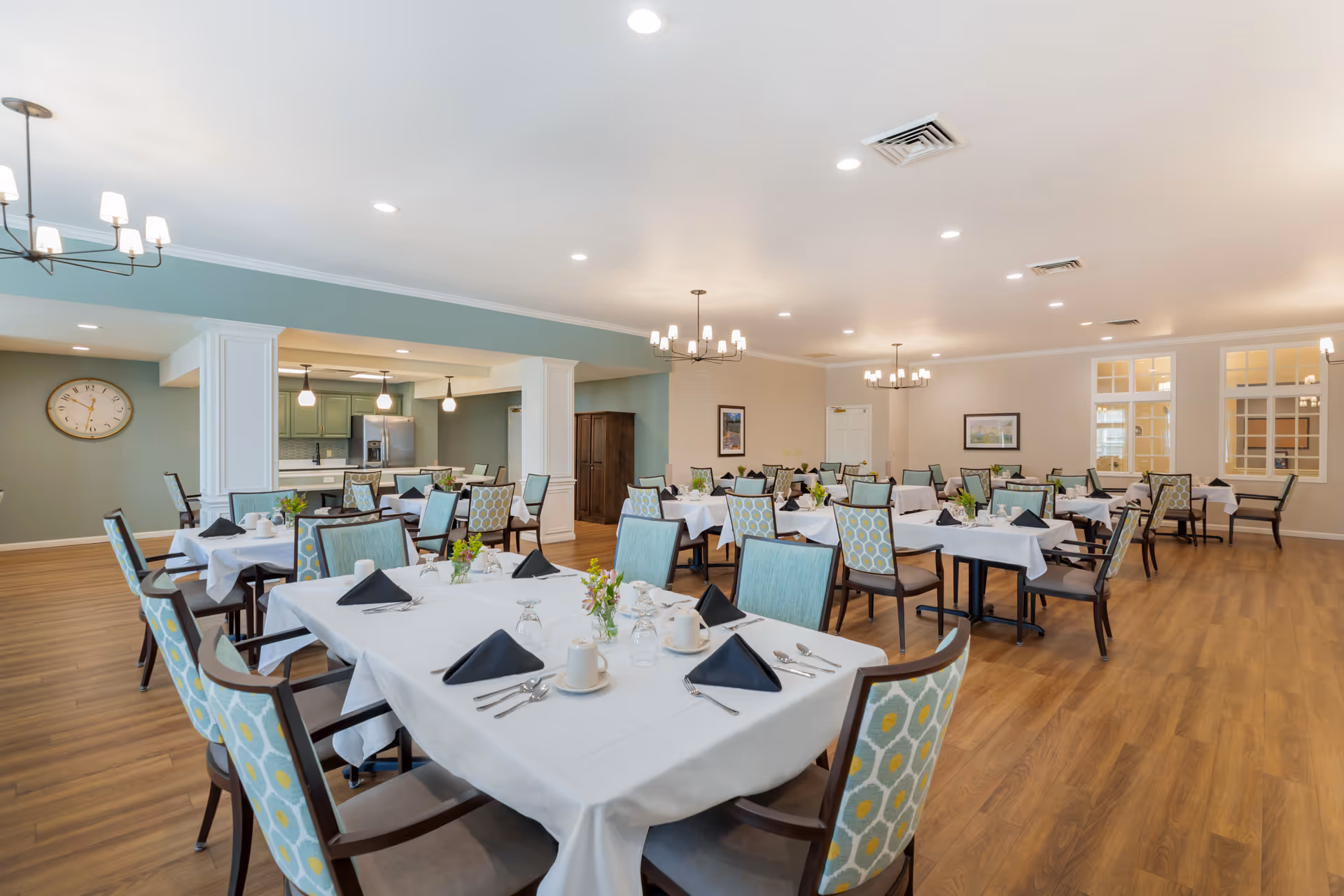 A spacious dining room with multiple tables covered in white tablecloths, each set with black folded napkins, cups, glasses, and silverware. The chairs have light blue upholstery with a yellow and white pattern on the backrests. The room features wooden flooring, soft lighting from chandeliers and recessed ceiling lights, and a wall clock. In the background, there is a kitchen area with green cabinetry and stainless steel appliances.