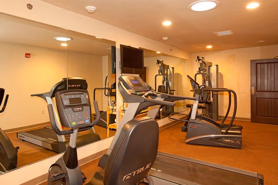 Small fitness room with exercise equipment including a treadmill, stationary bike, and elliptical machine. The room has a large mirror on one wall, a mounted TV, and a wooden door. The floor is carpeted and the walls are painted beige.