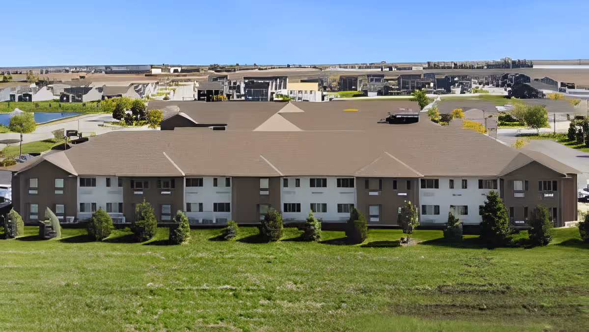 Exterior view of a two-story senior living facility building with a brown roof and white and brown walls, surrounded by green grass and small trees under a clear blue sky.