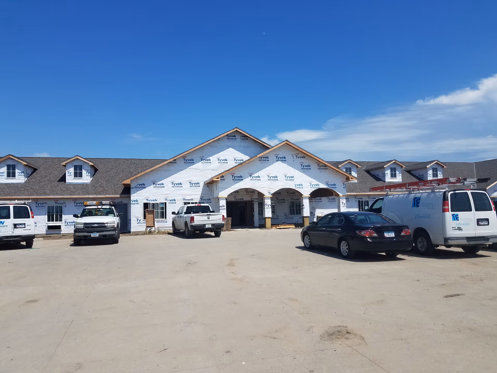 Front exterior view of a building under construction with Tyvek house wrap visible on the walls. Several vehicles, including trucks and vans, are parked in front of the building under a clear blue sky.