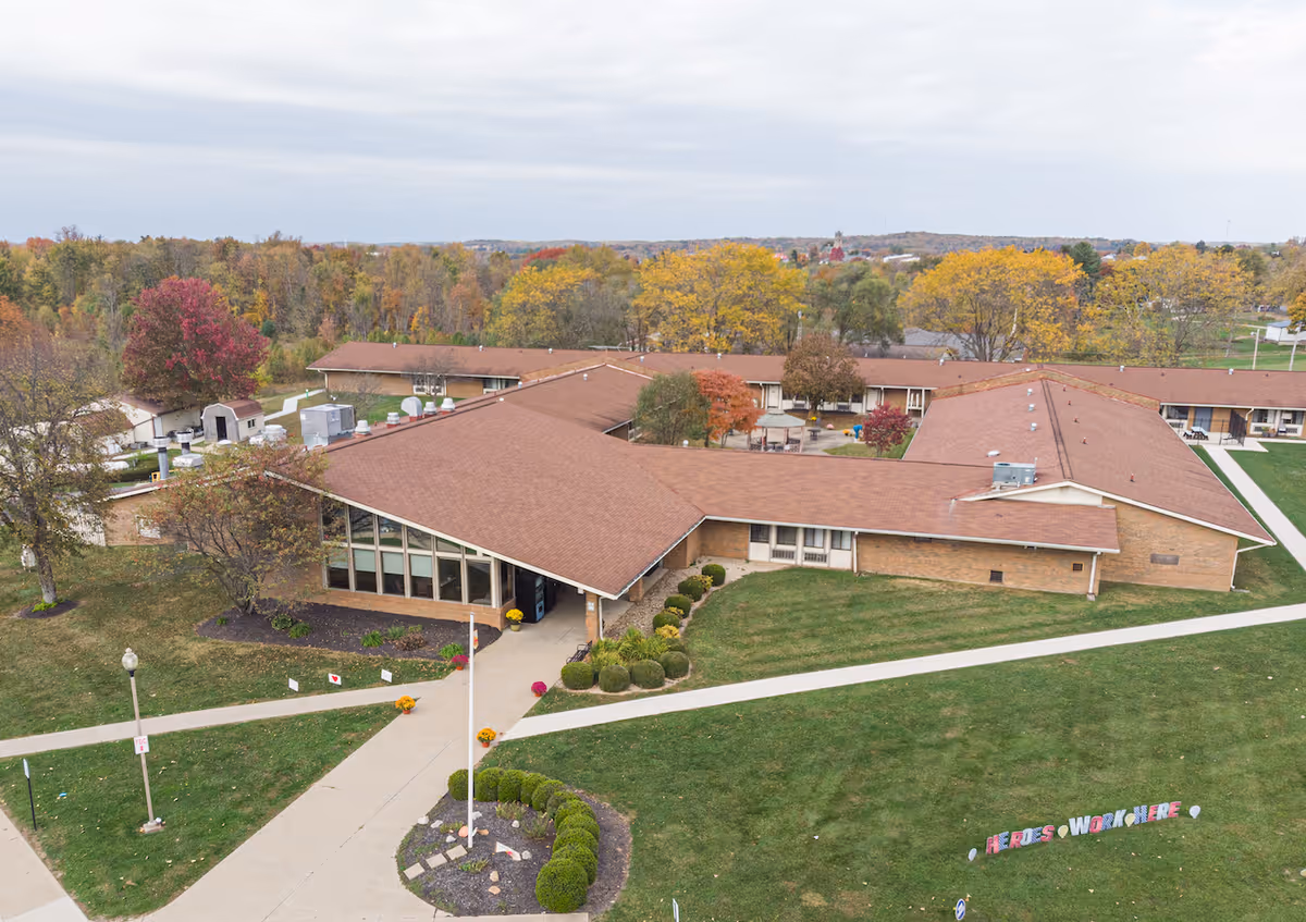Aerial view of a single-story senior living facility building with a brown roof surrounded by green lawns and trees with autumn foliage. There are paved walkways leading to the entrance, and a sign on the lawn reads 'HEROES WORK HERE'.