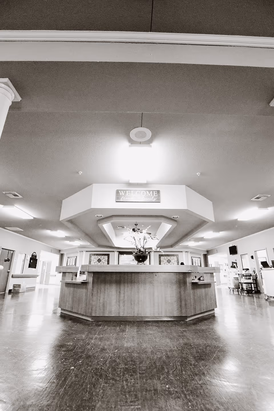 Interior view of a healthcare facility reception area with a large wooden reception desk in the center, a flower arrangement on top, and a 'WELCOME' sign hanging above. The space is well-lit with ceiling lights and has a polished floor.