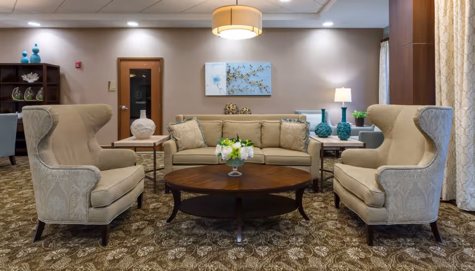 A cozy living room area with two beige wingback chairs facing a beige sofa. A round wooden coffee table with a flower arrangement is in the center. The room has patterned carpet, side tables with decorative vases, a lamp, and wall art. Light beige walls and curtains complete the warm and inviting atmosphere.
