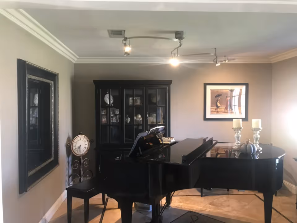 Interior room featuring a black grand piano with sheet music on the stand, a black cabinet with glass doors displaying decorative items, a large wall mirror, a standing clock, and a framed artwork on the wall. The room has beige walls, tiled floor, and ceiling track lighting.