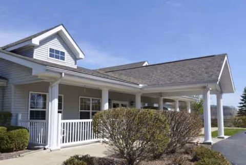 White senior living facility exterior with porch, columns, and trimmed hedges under blue sky