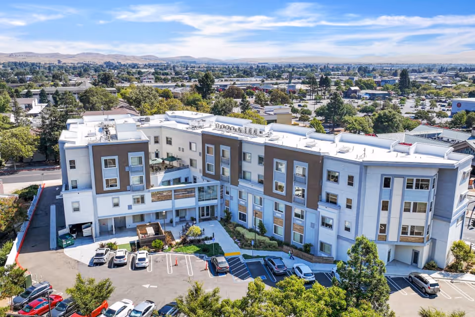 Aerial view of a modern three-story senior living facility building with white and brown exterior walls, surrounded by trees and parked cars. The building has multiple windows and a small courtyard area with greenery. The background shows a suburban area with more trees, buildings, and distant hills under a partly cloudy sky.