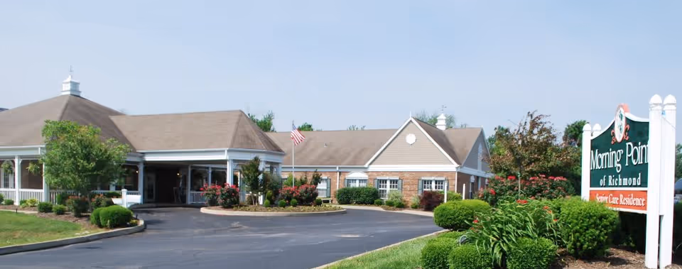 Exterior view of Morning Pointe of Richmond senior care residence showing a single-story building with a covered entrance, surrounded by well-maintained landscaping including bushes, flowers, and trees, with an American flag near the entrance and a clear sky above.
