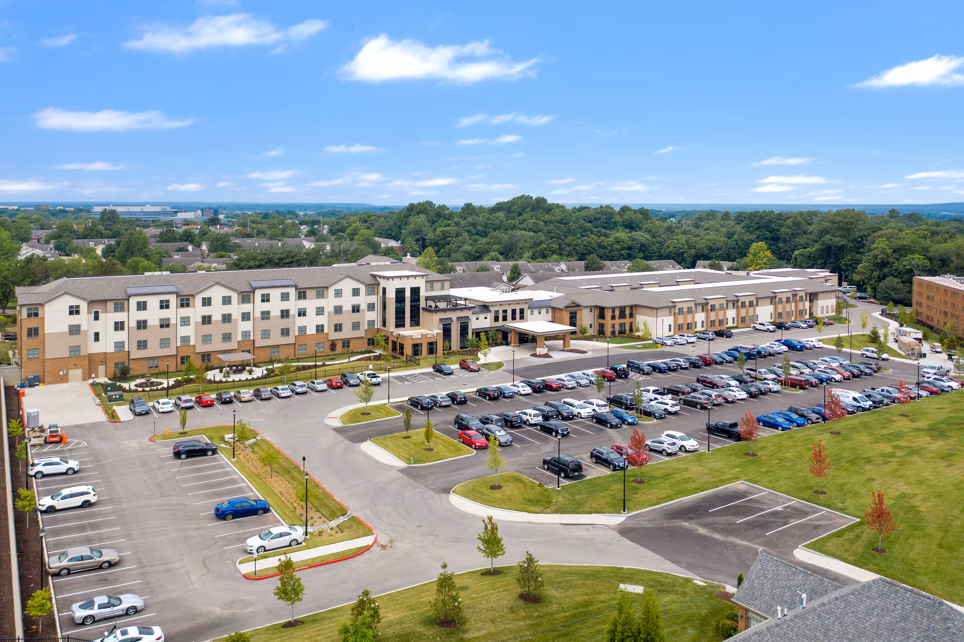 Aerial view of Friendship Village Chesterfield, showing a large senior living facility building with multiple floors, surrounded by a spacious parking lot filled with cars. The building is set against a backdrop of trees and a partly cloudy blue sky.