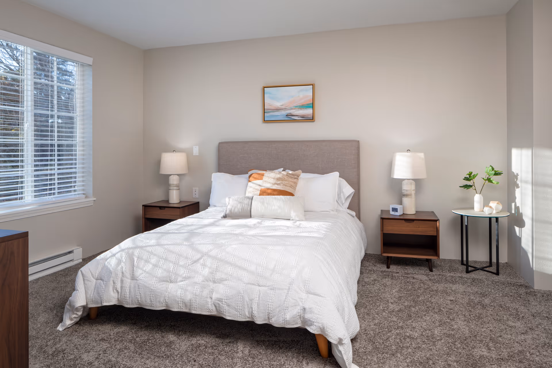 A neatly made bed with white bedding and decorative pillows in a bedroom. The bed has a gray upholstered headboard and is flanked by two wooden nightstands, each with a white lamp. There is a small round table with a plant and decorative items on the right side of the bed. A window with white blinds is on the left wall, and a framed landscape painting hangs above the bed. The room has beige walls and carpeted flooring.