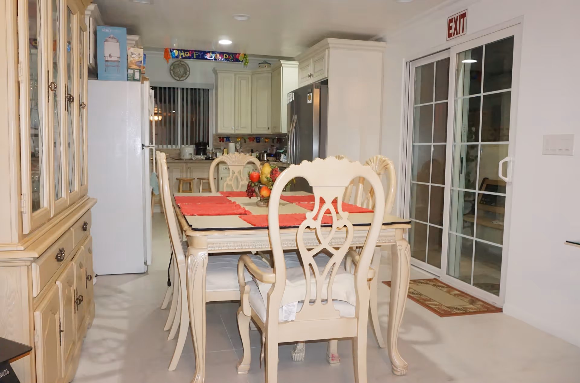 Interior view of a dining area with a cream-colored dining table and six matching chairs with cushions. The table is set with red placemats and a fruit centerpiece. In the background, there is a kitchen with white cabinets, a refrigerator, and various kitchen items on the counters. A sliding glass door with an exit sign above it is visible on the right side of the image.
