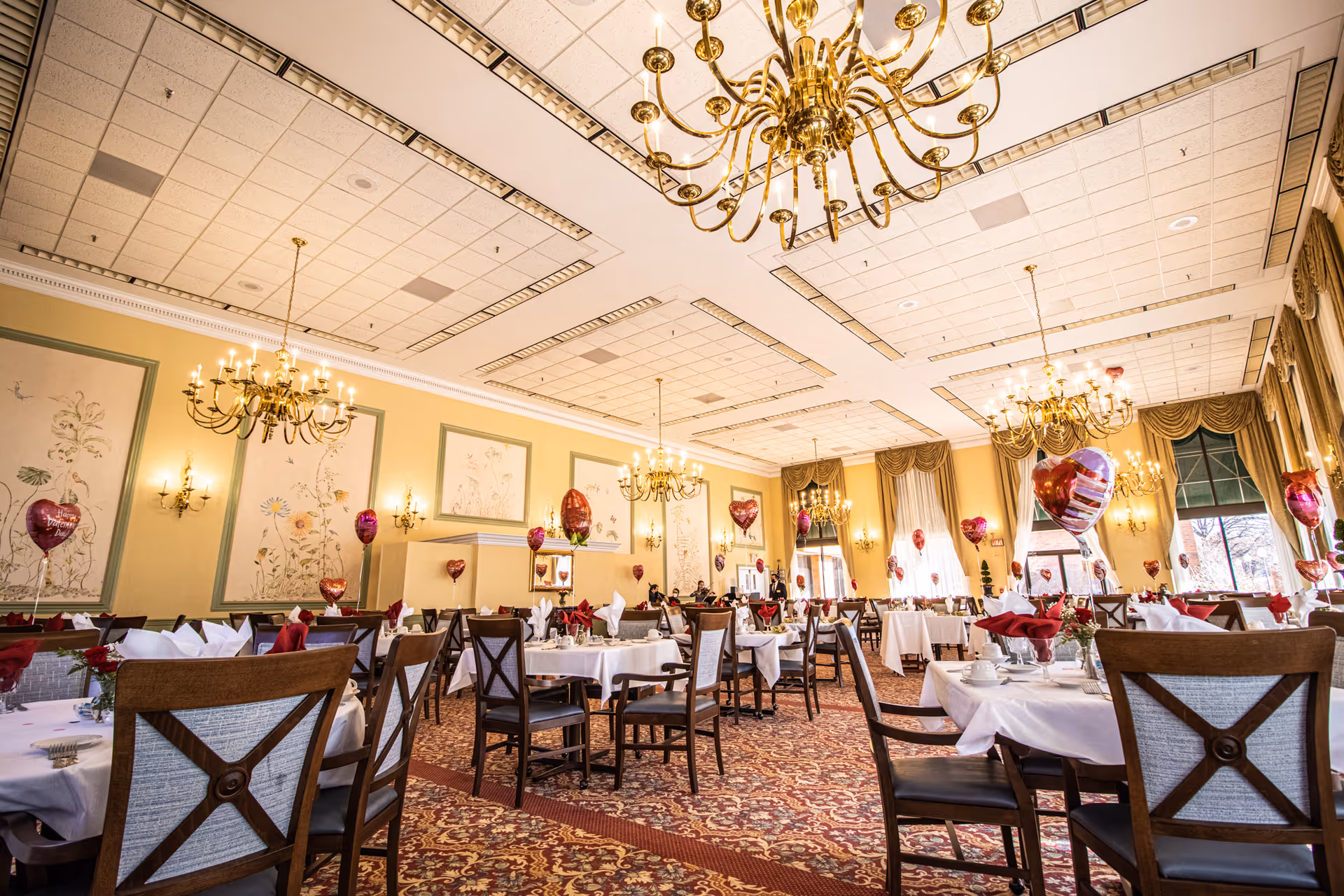 A spacious and elegant dining room decorated with chandeliers, wall sconces, and large windows with drapes. The tables are set with white tablecloths, red napkins, and heart-shaped balloons, creating a festive atmosphere.