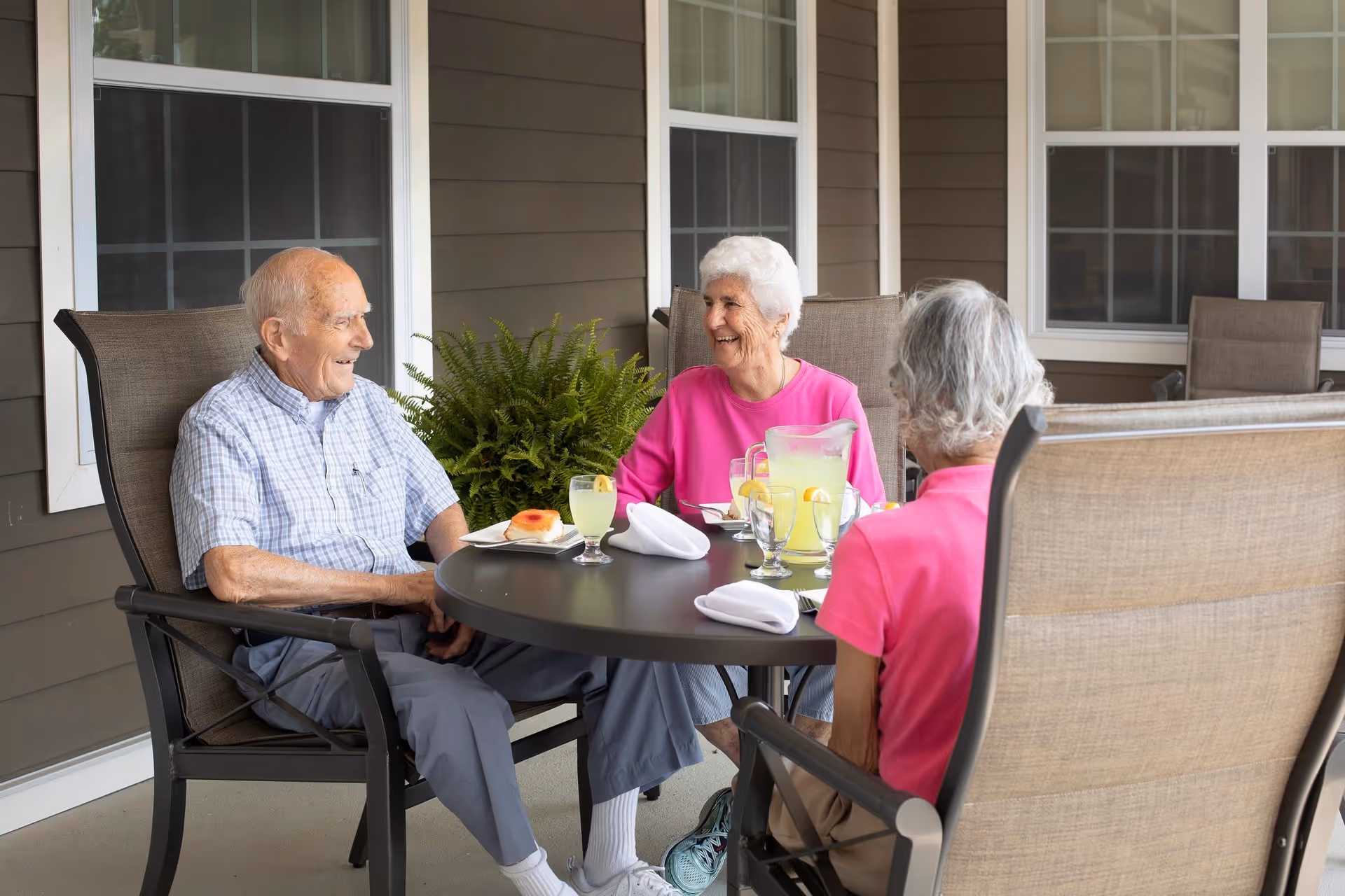 Three elderly people seated around a round patio table on a covered porch, smiling and sharing lemonade.