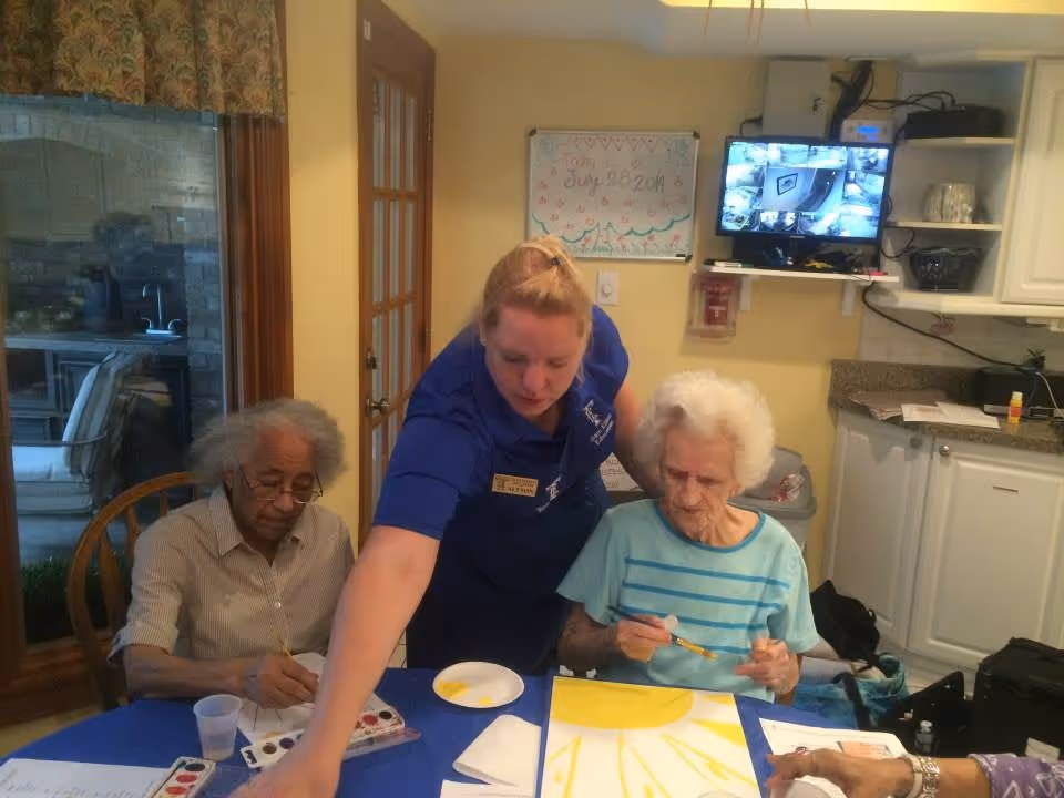 A caregiver in a blue uniform assists two elderly women seated at a table covered with a blue tablecloth. They are engaged in painting activities with watercolors and brushes. The room has yellow walls, a whiteboard with the date July 28, 2014, and a monitor displaying security camera footage mounted on the wall. The setting appears to be a common area or activity room in a senior living facility.