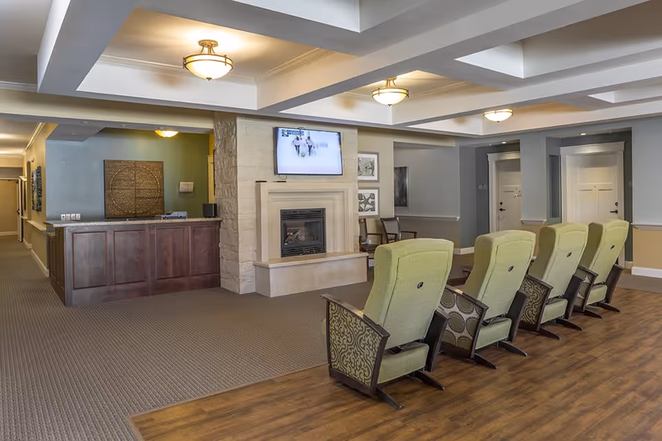 A cozy common area in a senior living facility featuring a row of green upholstered reclining chairs facing a wall-mounted TV above a fireplace. The room has a mix of carpet and wood flooring, a wooden reception desk, and soft ceiling lighting with recessed panels.