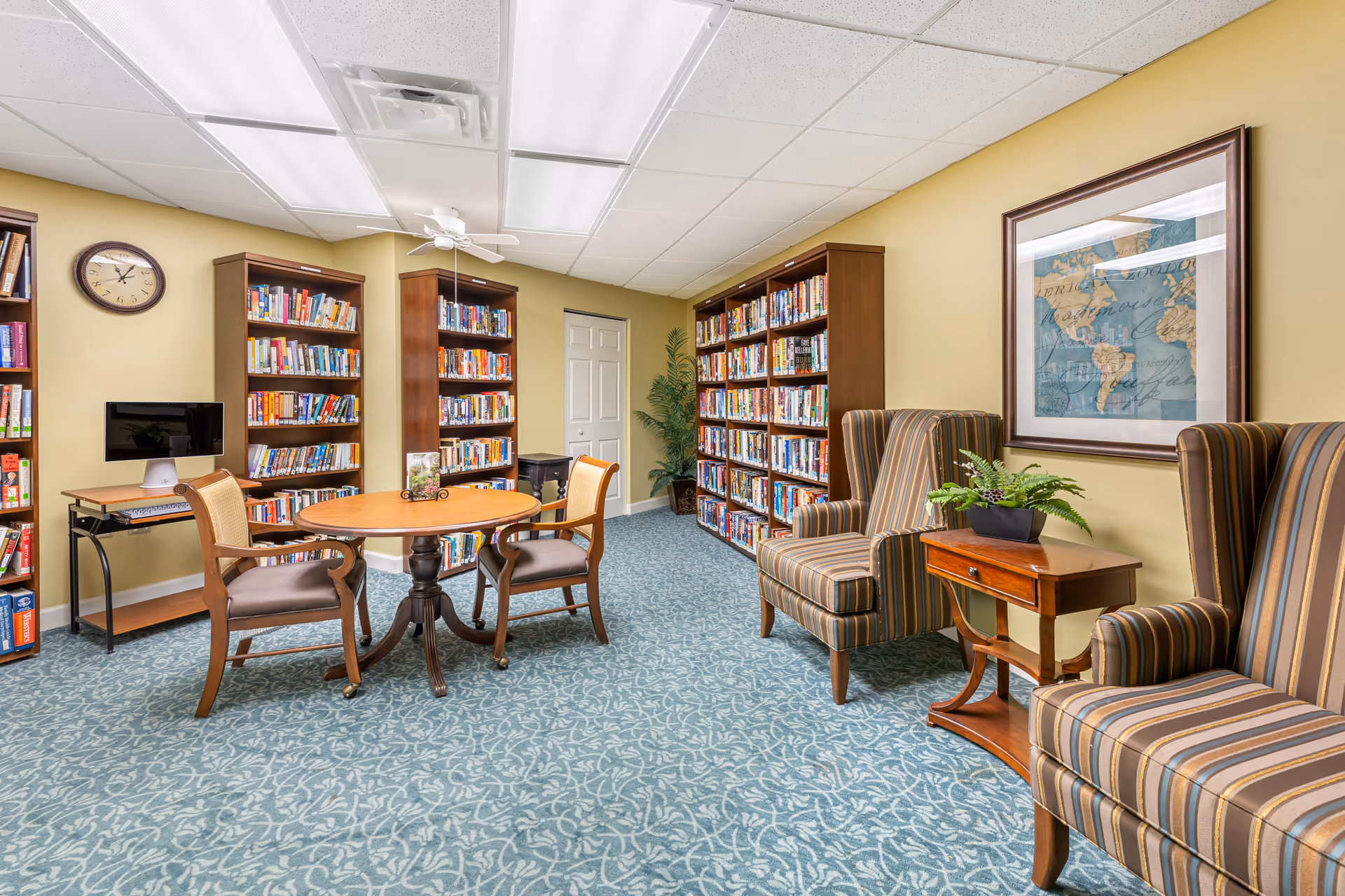 A cozy library room with multiple wooden bookshelves filled with books, a round wooden table with two chairs, two striped armchairs separated by a small wooden table with a plant, a wall clock, a computer on a small desk, and a framed world map on the wall. The room has a patterned blue carpet and yellow walls.