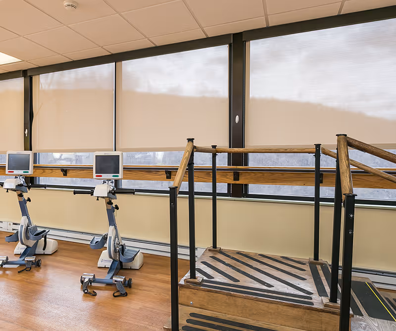 Physical therapy room with two exercise bikes and a wooden parallel-bar walking ramp in front of large windows with roller shades.