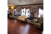Sunlit common room with upholstered chairs and small tables arranged along windows with decorative valances and hardwood flooring.
