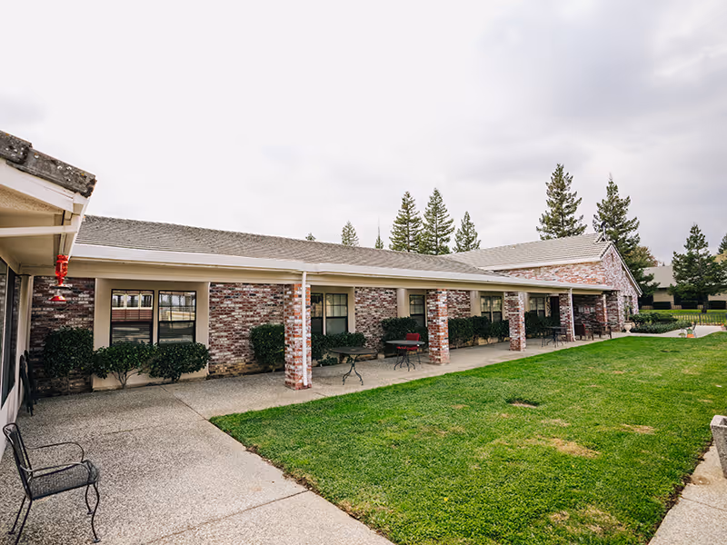 Single-story brick assisted living building with a covered patio, columns, patio tables and a grassy courtyard.