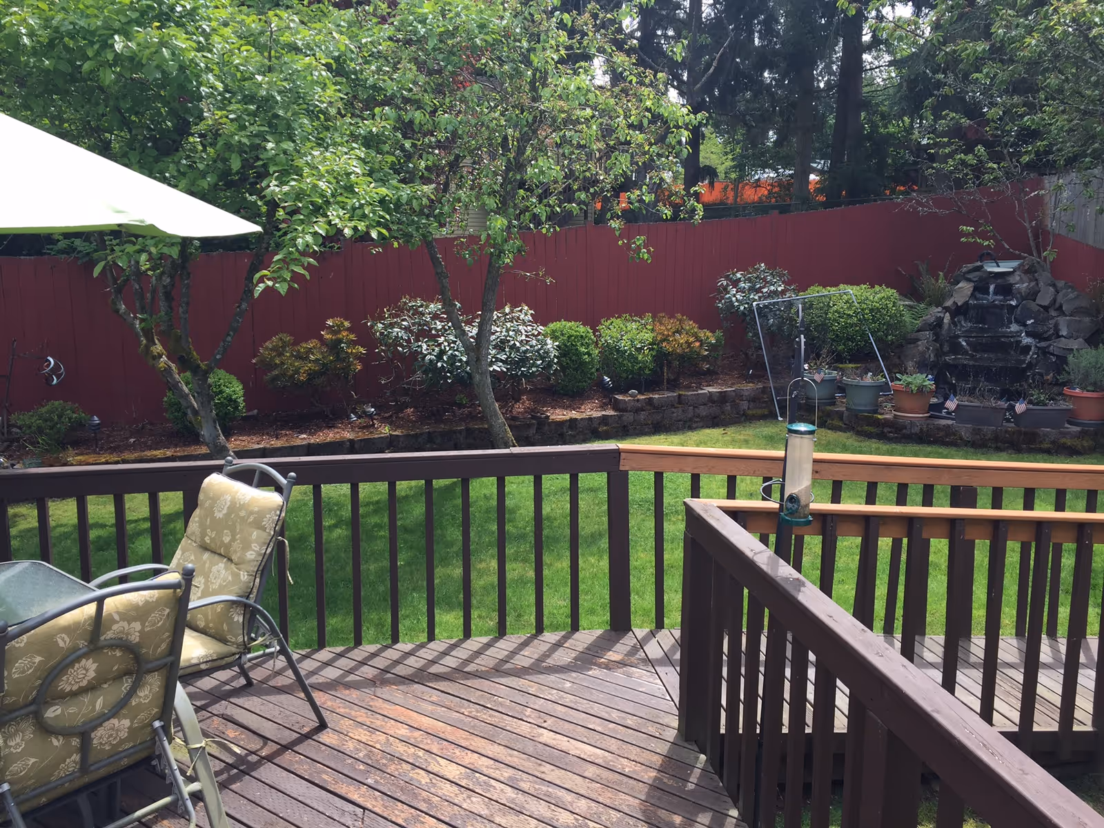 A wooden deck with patio furniture including cushioned chairs and a glass table, overlooking a green lawn with trees, shrubs, and a red wooden fence. There is a small rock waterfall feature with potted plants near the fence.
