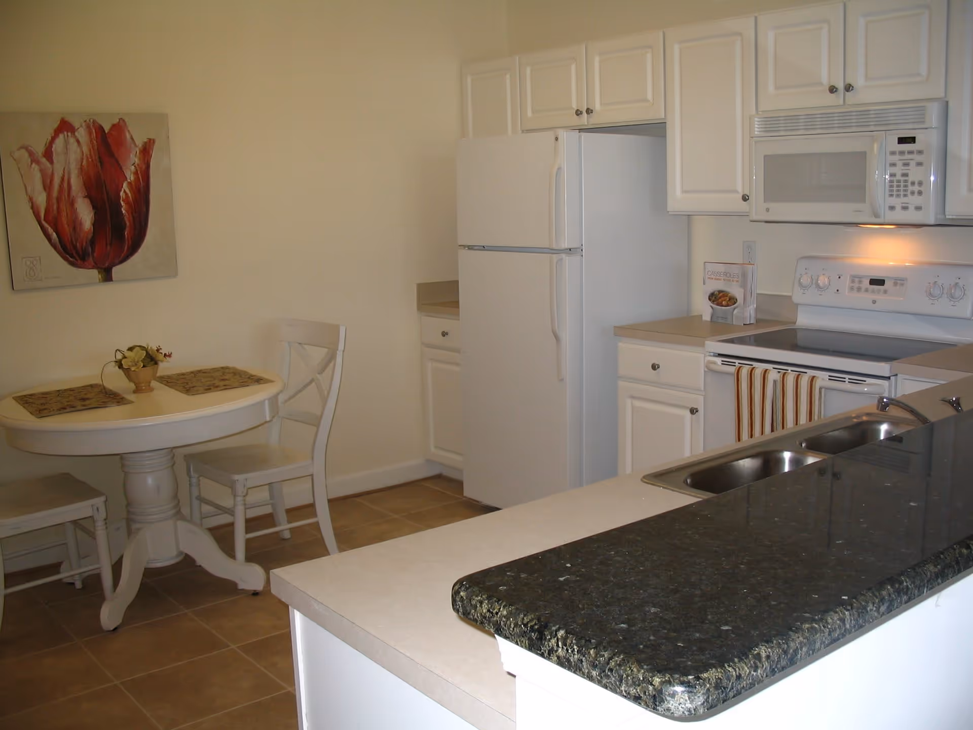 A kitchen area with white cabinets, a white refrigerator, a white microwave above a white stove, and a double sink with a dark granite countertop. To the left, there is a small round dining table with two chairs, a small plant centerpiece, and a large painting of a red flower on the wall.