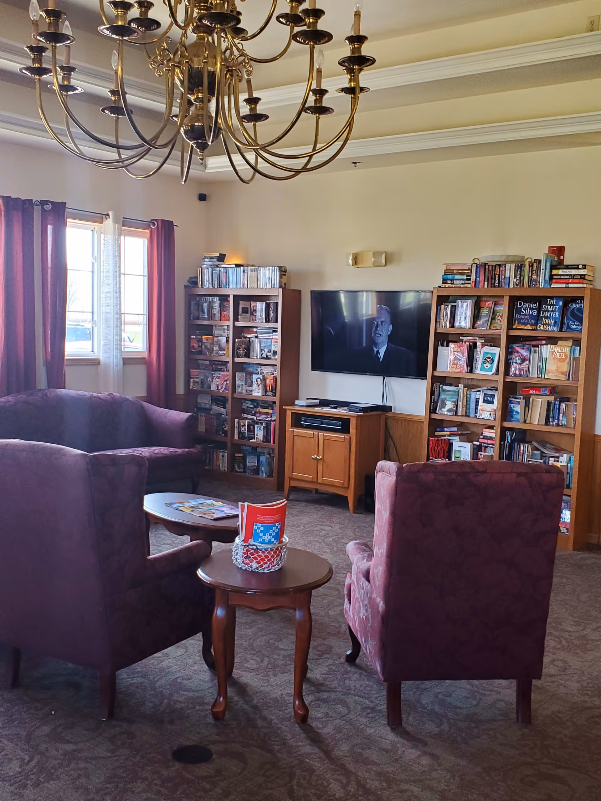 A cozy living room area with two purple armchairs and a matching loveseat arranged around two wooden coffee tables. The room has a large window with purple curtains letting in natural light. There are two wooden bookshelves filled with books and DVDs, and a flat-screen TV mounted on the wall showing a black and white image of a man. A brass chandelier hangs from the ceiling.