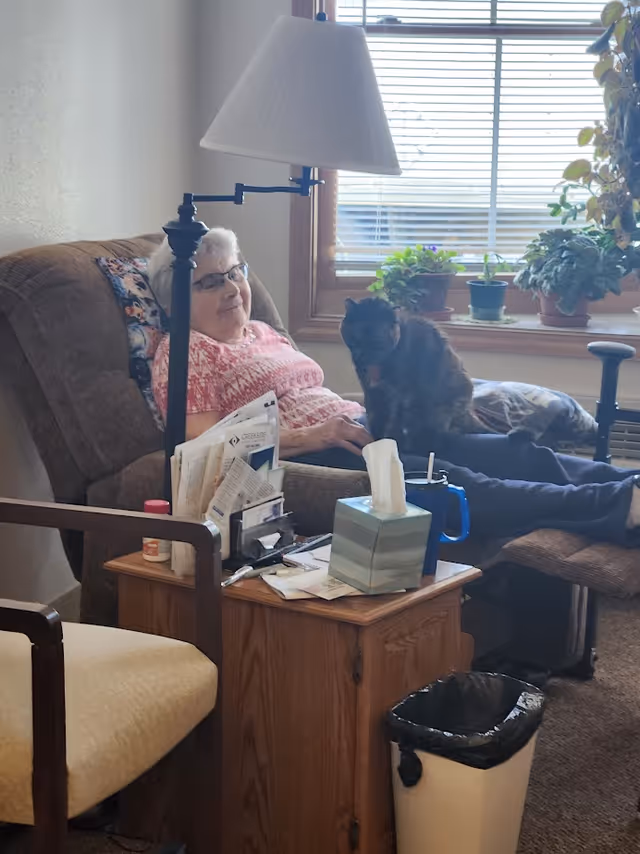 An elderly woman wearing glasses and a pink patterned shirt is sitting comfortably in a brown recliner chair with a cat on her lap. There is a wooden side table next to her with tissues, papers, and a blue cup. A lamp is positioned behind the chair, and a window with several potted plants on the windowsill is in the background. A beige cushioned chair and a trash bin are also visible in the room.