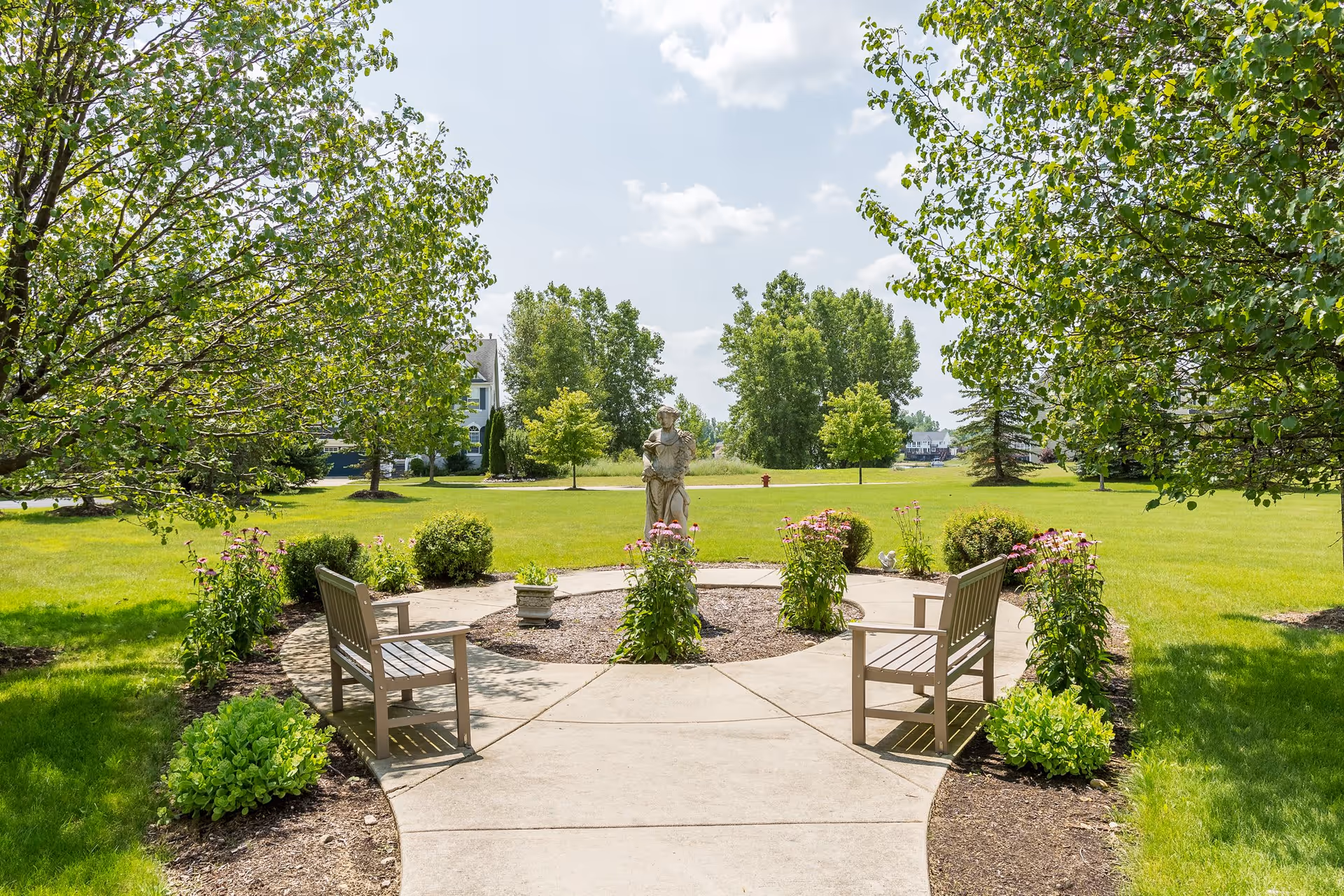 A peaceful outdoor garden area with a circular concrete pathway surrounding a statue in the center. Two wooden benches face the statue, and the garden is bordered by green bushes and flowering plants. Large trees and a grassy lawn extend into the background under a partly cloudy sky.