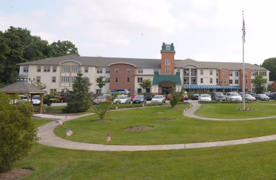 Front exterior view of Bristol Glen, a UMC Community, showing a three-story building with a green roofed entrance, a clock tower, multiple windows, a parking lot with cars, a gazebo on the left, a flagpole with an American flag, and a well-maintained lawn with small American flags along the curved walkway.
