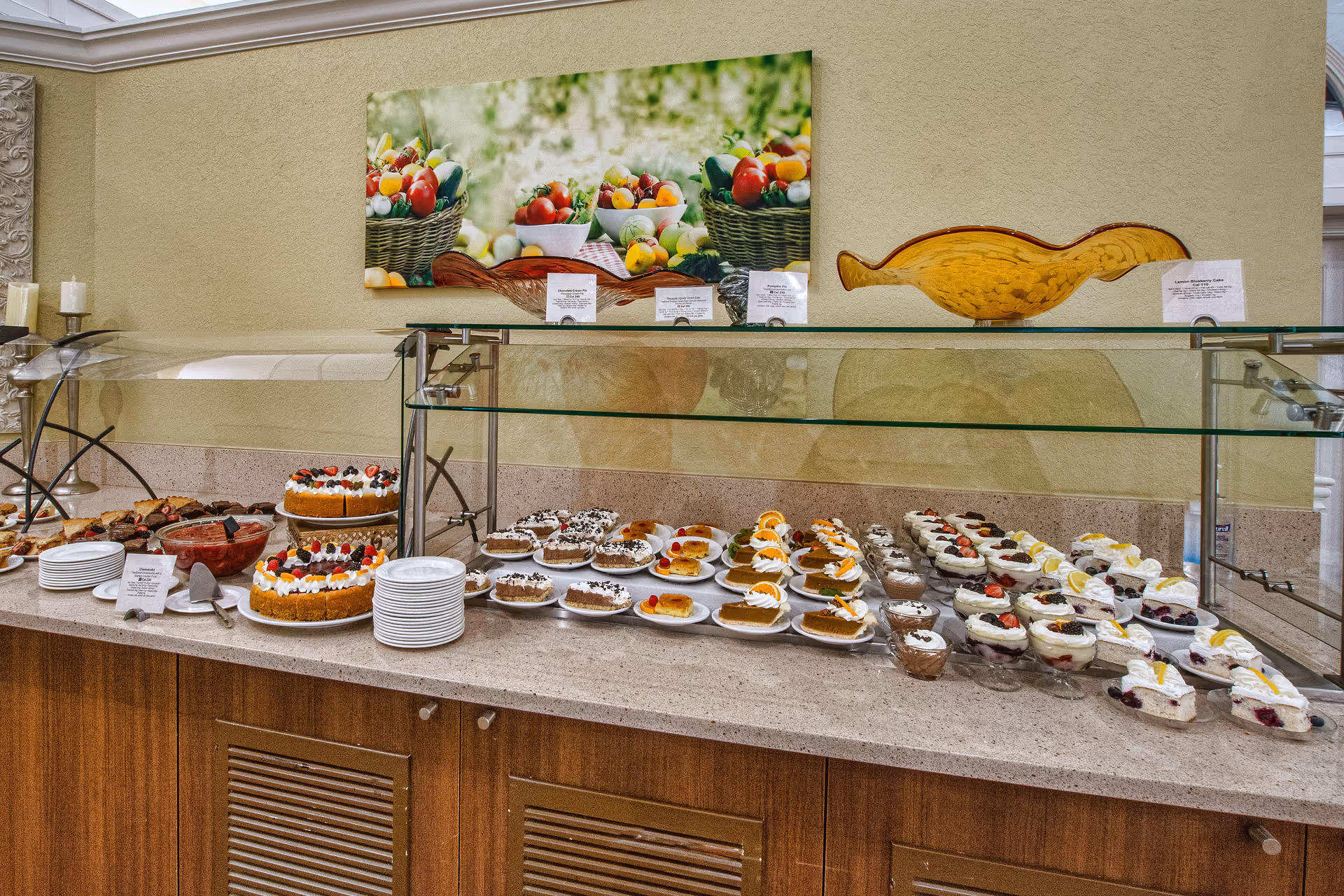 A dessert buffet with various cakes, pies, and parfaits arranged on a countertop under a glass sneeze guard. Plates and serving utensils are also visible. Above the buffet is a colorful painting of baskets filled with fruits and vegetables.
