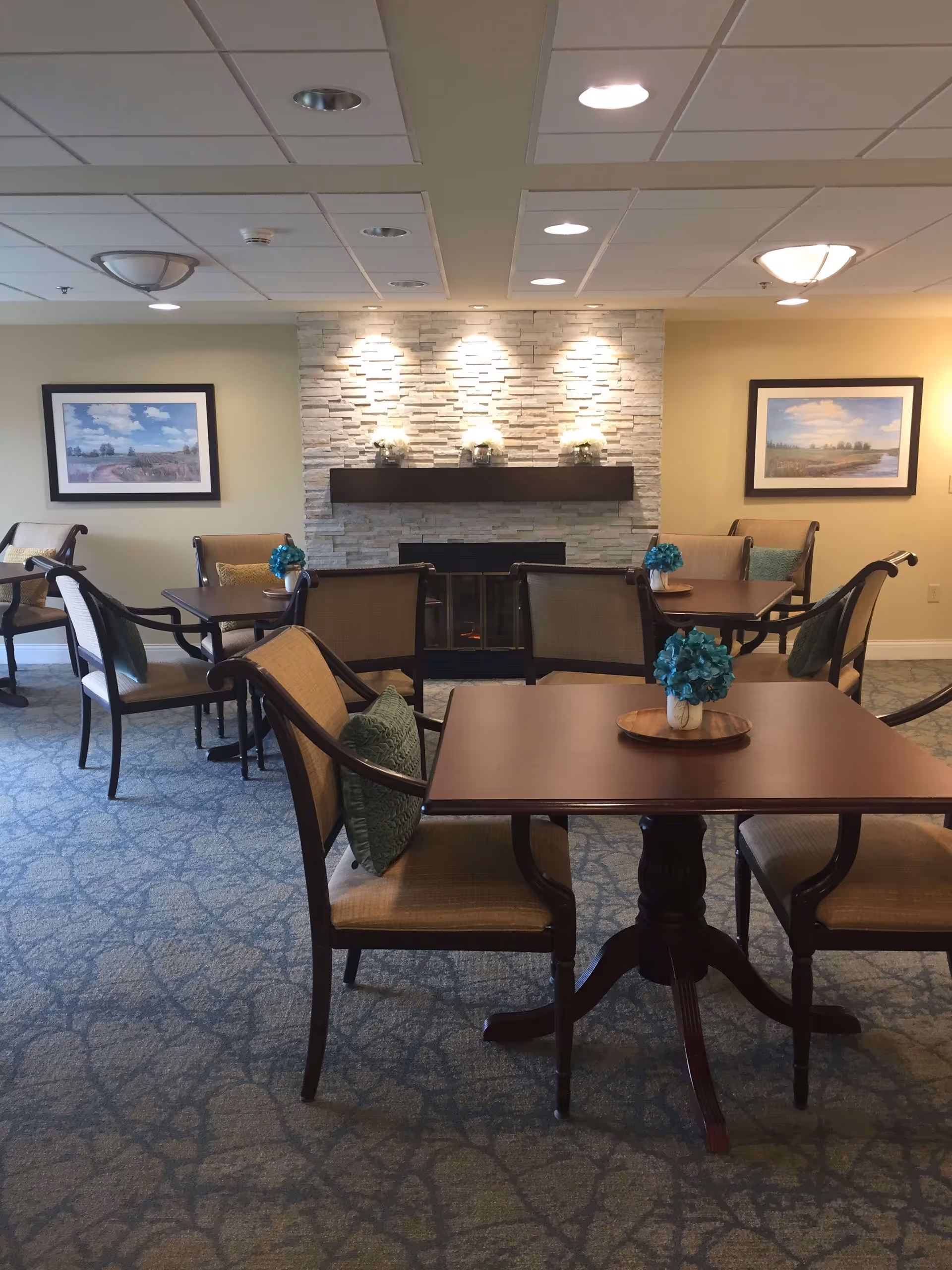 A cozy common area with several wooden tables and cushioned chairs arranged on a patterned carpet. Each table has a small vase with blue flowers. The room features a stone fireplace with a wooden mantel decorated with small flower arrangements. Two framed landscape paintings hang on the yellow walls on either side of the fireplace. The ceiling has recessed lighting and two ceiling light fixtures.
