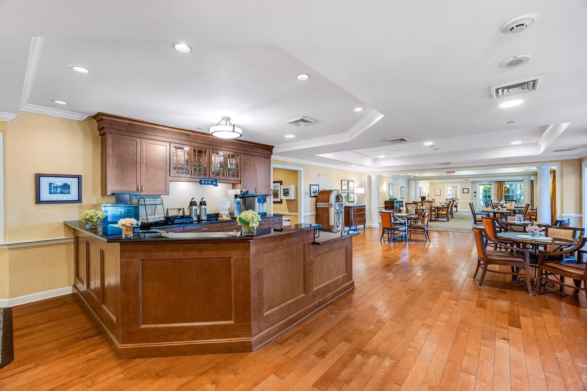 Spacious interior of a senior living facility featuring a wooden bar area with coffee machines and glass cabinets, adjacent to a dining area with multiple tables and chairs arranged for residents. The room has hardwood floors, recessed lighting, and large windows allowing natural light.