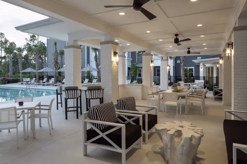 Covered outdoor seating area with modern chairs and tables next to a swimming pool. The area features ceiling fans, white brick columns, and ambient lighting. In the background, there are poolside lounge chairs with striped umbrellas and palm trees.