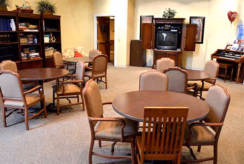 A senior common lounge with round wooden tables and upholstered chairs, a TV in a cabinet, bookshelves and an organ.