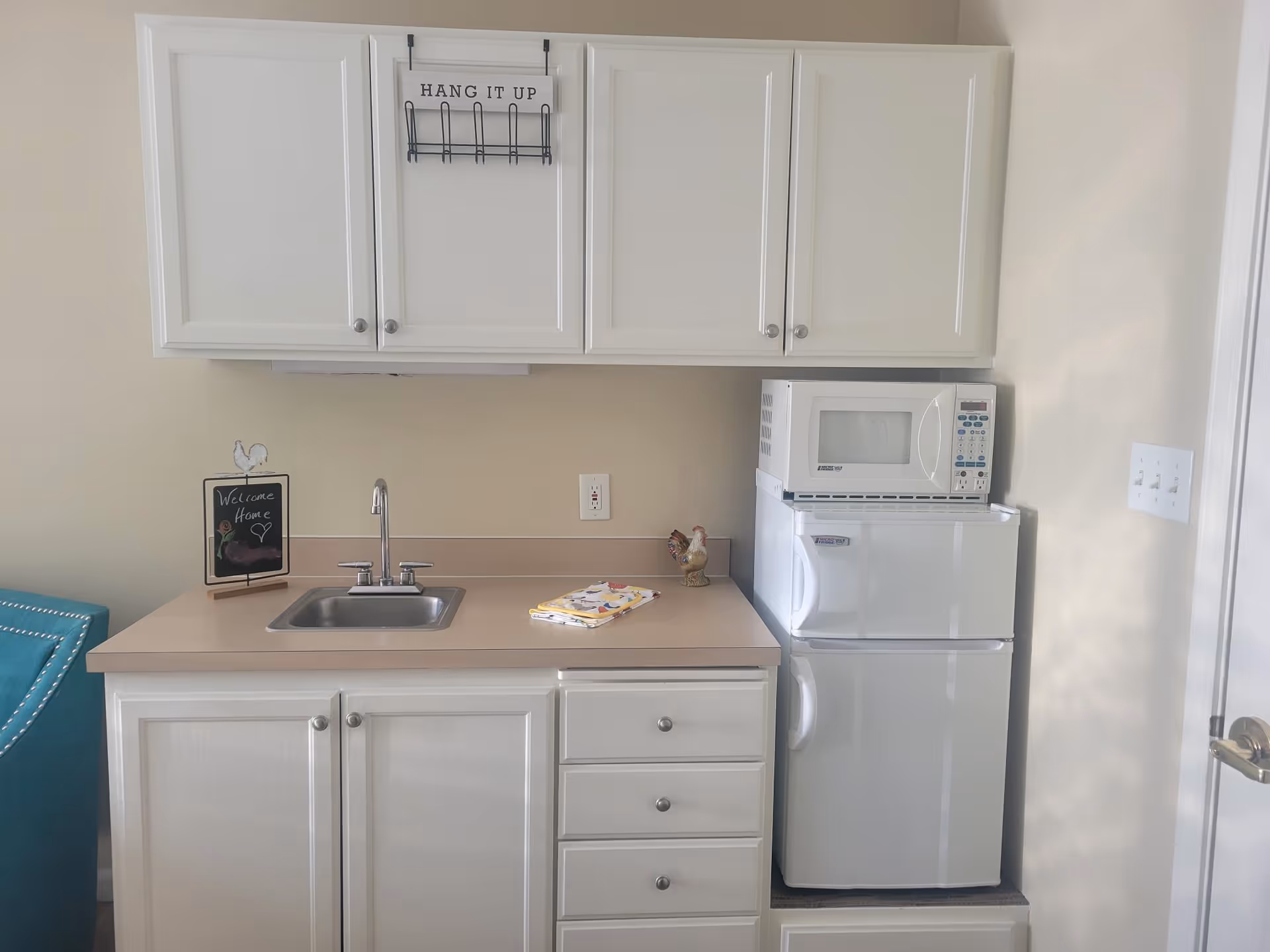Small kitchenette with white cabinets, a sink on a beige countertop, a microwave on a mini refrigerator, and small decorative items.