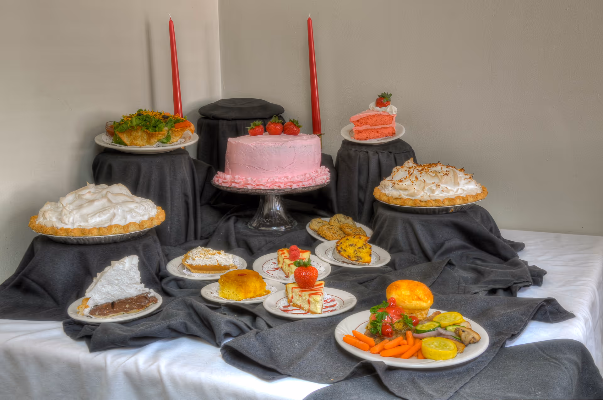 A display of various desserts and a plate of vegetables with a biscuit arranged on a table covered with a white tablecloth and dark gray fabric. The desserts include a pink frosted cake with strawberries on top, slices of pink cake, pies with meringue topping, a slice of chocolate pie, small cakes with strawberries, and other assorted pastries. Two red candles are placed behind the pink cake.