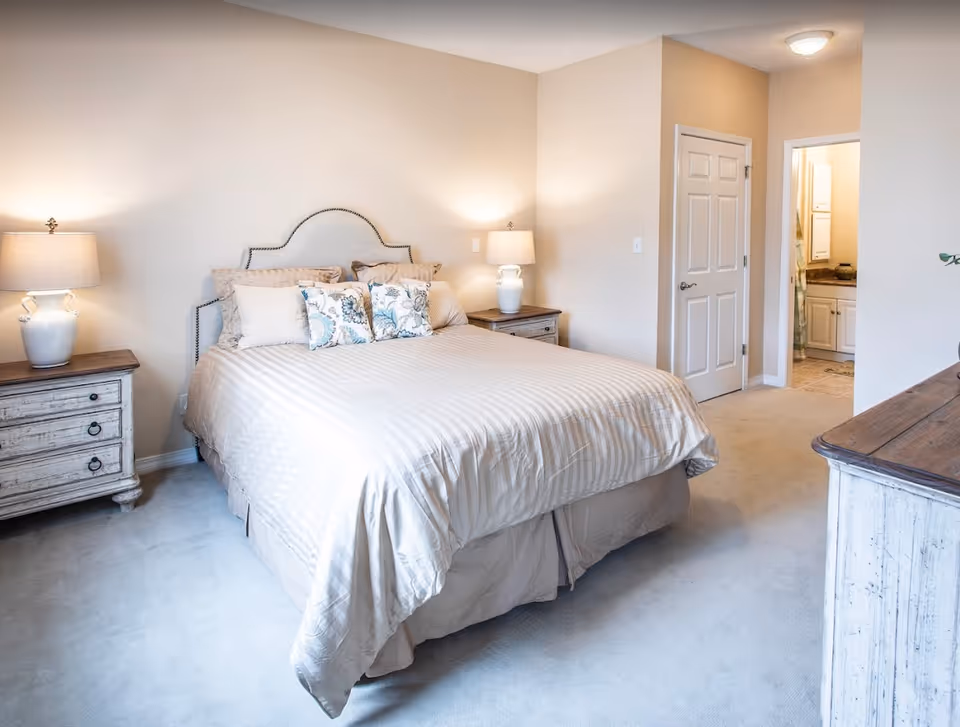 Bright, neutral-toned bedroom with a bed, two bedside tables with lamps, and a doorway leading to a bathroom.