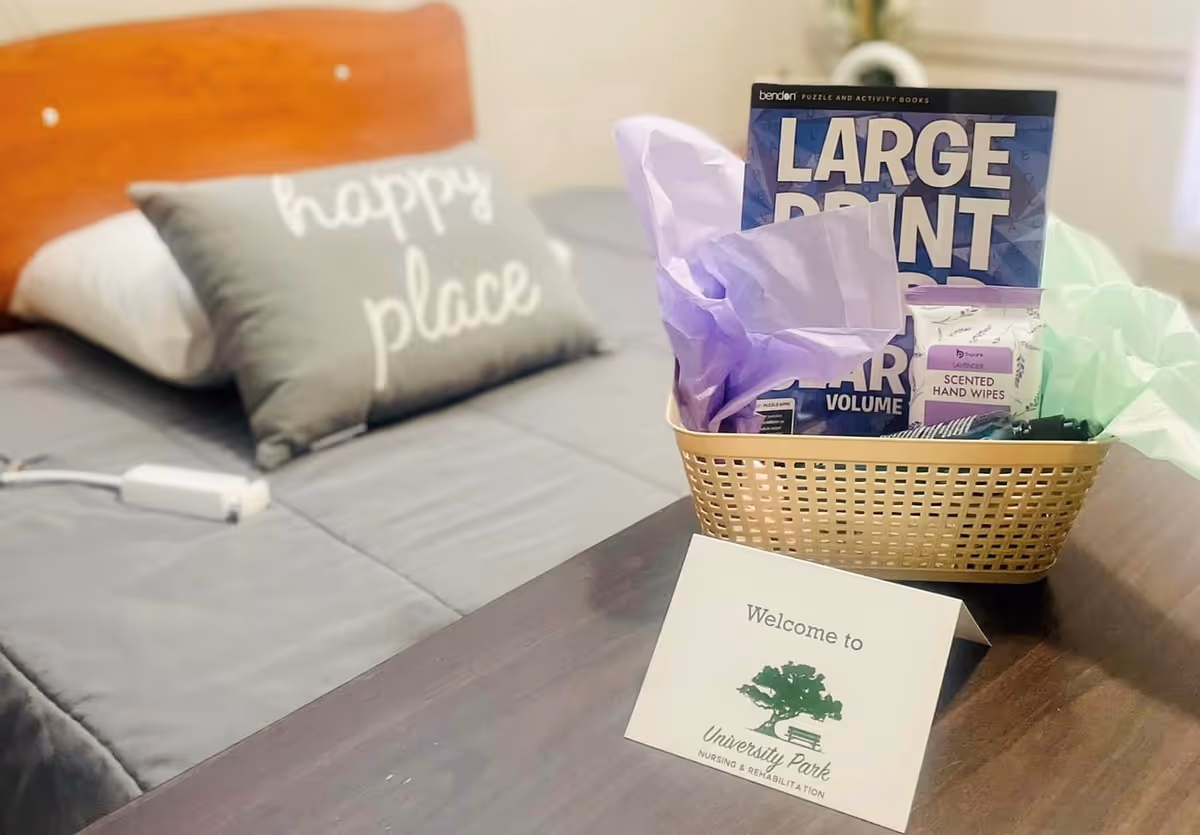A neatly made bed with a gray pillow that says 'happy place' and a gray blanket. On a nearby table, there is a basket containing a large print puzzle book, scented hand wipes, and purple tissue paper. In front of the basket, there is a card that reads 'Welcome to University Park Nursing & Rehabilitation' with a green tree logo.