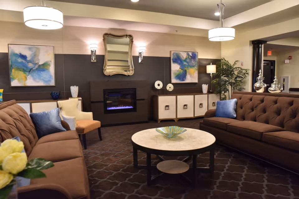 Comfortable lounge with two brown tufted sofas facing a round coffee table, a wall-mounted electric fireplace, artwork, and pendant lights.