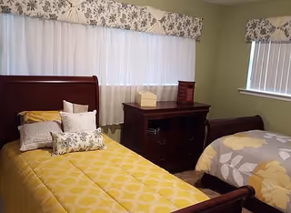 Two twin sleigh beds with yellow and gray bedding flank a wooden dresser beneath a curtained window in a softly painted bedroom.