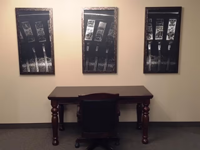 A dark wooden desk with a black office chair in front of it is positioned against a beige wall. Above the desk, there are three framed black and white abstract art pieces arranged horizontally.