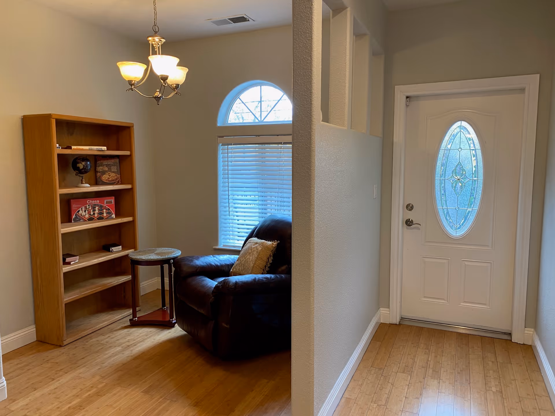 A cozy corner of a living area with a large brown leather armchair, a small round wooden side table, and a wooden bookshelf containing a globe, books, and a chess set. There is a window with blinds and an arched top letting in natural light. A white front door with an oval decorative glass panel is visible to the right, and a three-light chandelier hangs from the ceiling.