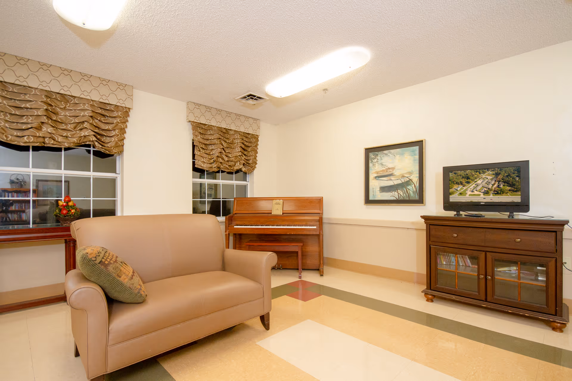 A cozy common area with a beige loveseat, a wooden piano against the wall, a TV on a wooden cabinet, and two windows with patterned valances. A framed painting hangs on the wall above the piano.