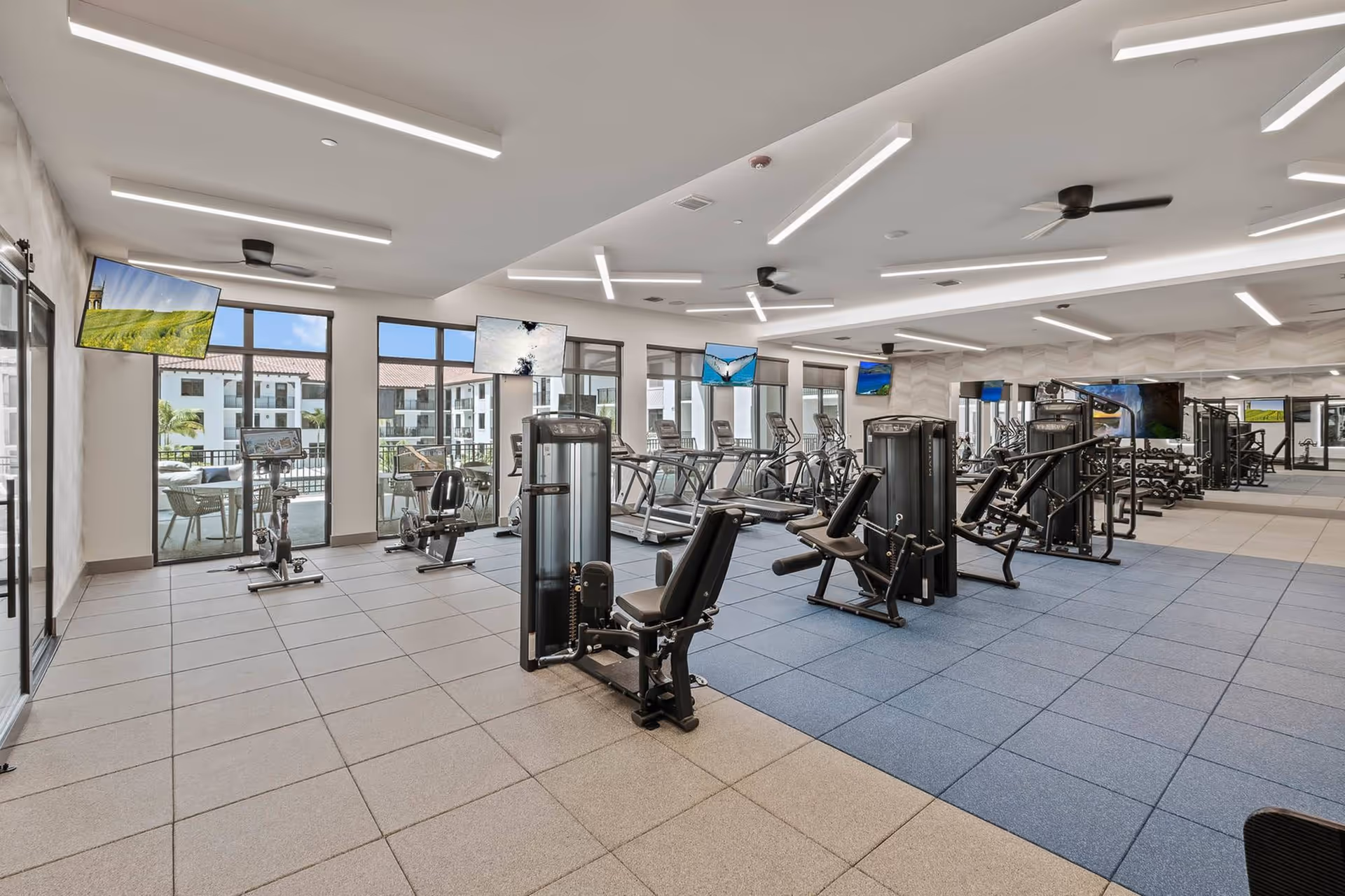 Bright modern gym with exercise machines, weight equipment, wall-mounted TVs, and large windows overlooking the complex.
