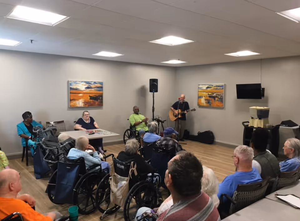 Residents in wheelchairs and chairs watch a musician playing guitar in a community activity room.