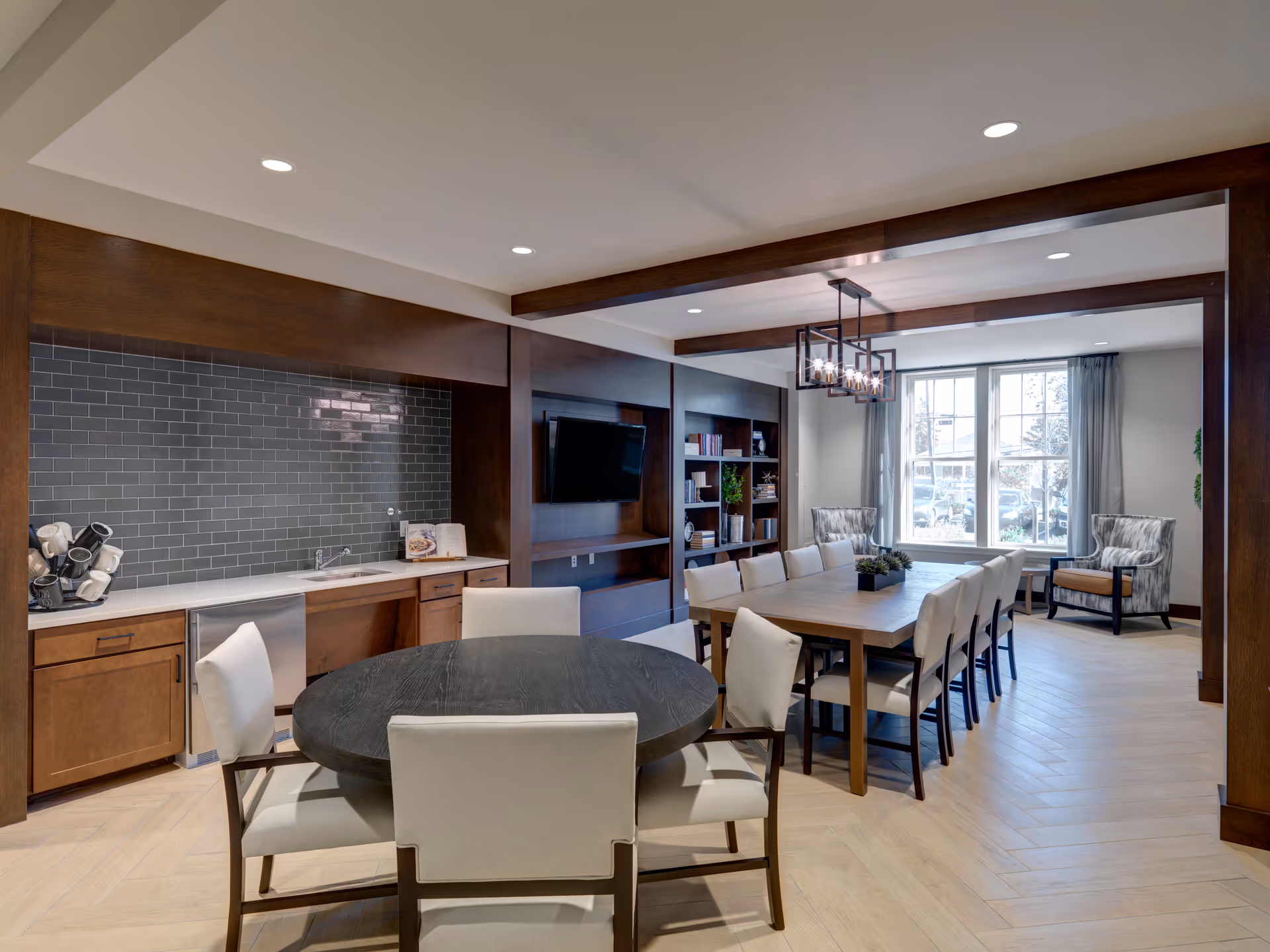 A spacious and well-lit common area featuring a round black table with four beige upholstered chairs in the foreground, and a long rectangular wooden dining table with twelve matching chairs in the background. The room has wooden cabinetry with a small sink and countertop along one wall, a mounted flat-screen TV, built-in bookshelves, large windows with gray curtains, and modern ceiling lights.